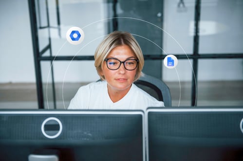 A woman wearing glasses works at a dual monitor work station