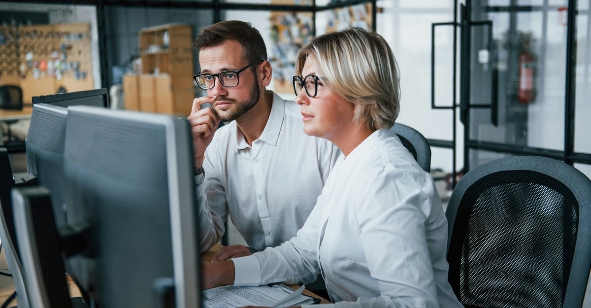 Two coworkers look at a desktop monitor together 